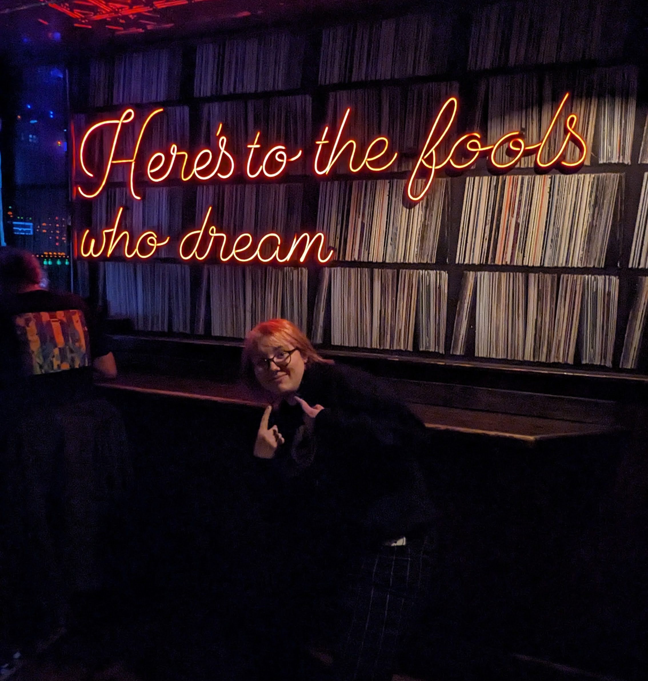 Nico giving a very snazzy thumbs up in front of a neon sign that says "Here's to the fools who dream."